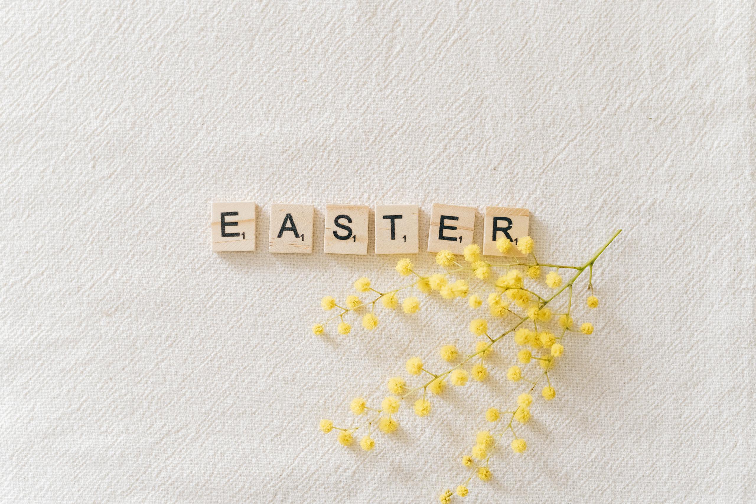 Flat lay of wooden letter tiles spelling Easter with yellow mimosa flower on textured background.